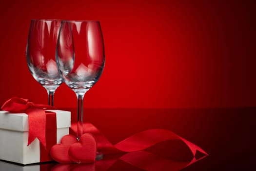 Gift box, red satin hearts and two empty wineglasses on a red background. Valentine's Day card with copy space. Design element for romantic greeting card, wedding invitation, Women's Day postcard