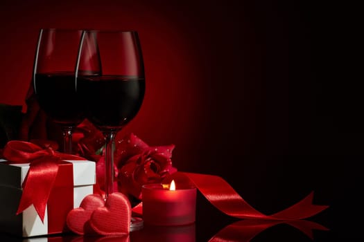 Still life with gift box, roses, red satin hearts and two glasses with wine on a red background. Valentine's Day card with copy space. Design element for romantic greeting card, wedding invitation