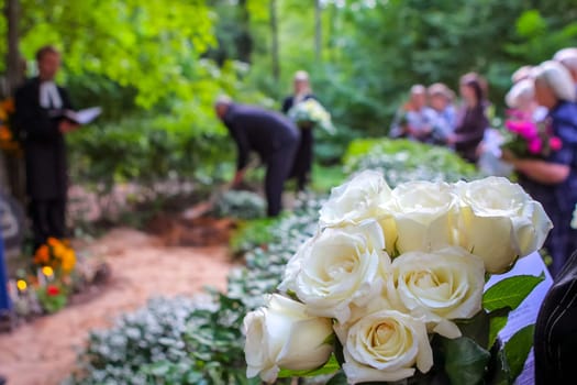 Roses in cemetery with people in the background. Funeral in cemetery   White roses in cemetery. Prayer at the tomb. Funeral ceremony. White roses at funeral near the grave.