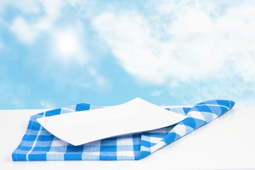 Empty plate background. Empty white plate on table with blue napkin or tablecloth over abstract blurred blue sky background. Template for your food and product display montage. Concept food. Copy space.