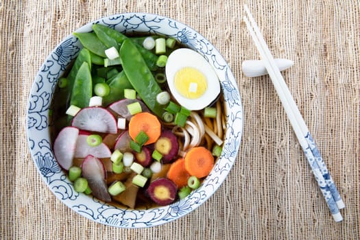 Filling bowl with udon noodle soup series final image: bowl with noodles, shiitake mushrooms, snow peas, broth, radishes, carrots, egg. and spring onions.