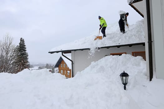 Two men shoveling high, heavy snow from a house roof