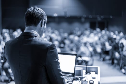 Speaker giving a talk on corporate business conference. Unrecognizable people in audience at conference hall. Business and Entrepreneurship event. Blue toned grayscale image.