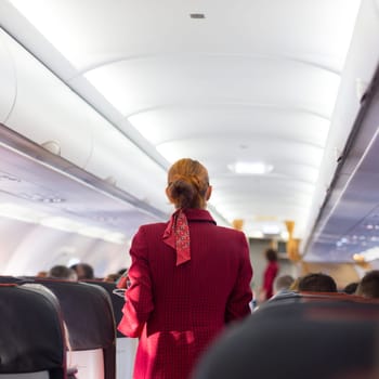 Stewardess in red uniform walking the aisle of commercial airplane. Unrecognizable passengers sitting on their seats during flight.