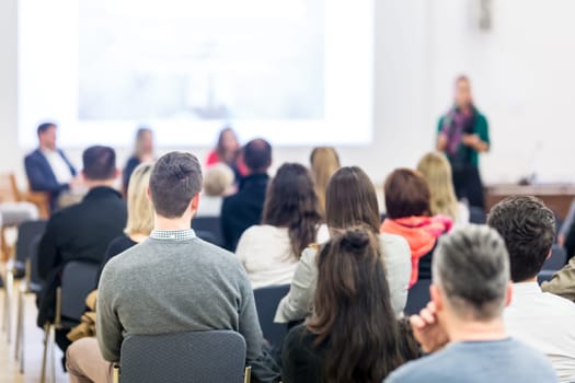 Business and entrepreneurship symposium. Female speaker giving a talk at business meeting. Audience in conference hall. Rear view of unrecognized participant in audience. Copy space on whitescreen.