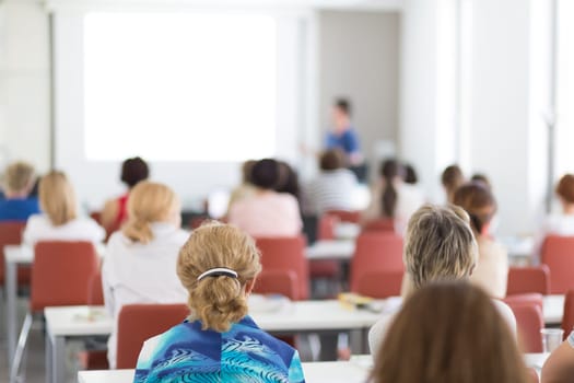 Female speaker giving presentation in lecture hall at university workshop. Audience in conference hall. Rear view of unrecognized participant in audience. Scientific conference event.