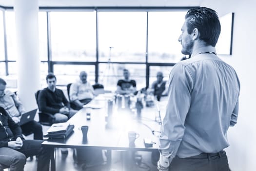Business man making a presentation at office. Business executive delivering a presentation to his colleagues during meeting or in-house business training. Rear view. Blue toned.