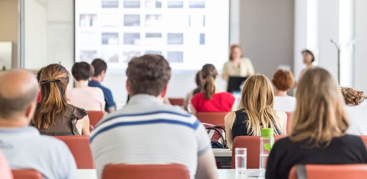 Female speaker giving presentation in lecture hall at university workshop. Audience in conference hall. Rear view of unrecognized participant in audience. Scientific conference event.