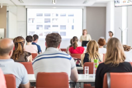 Female speaker giving presentation in lecture hall at university workshop. Audience in conference hall. Rear view of unrecognized participant in audience. Scientific conference event.