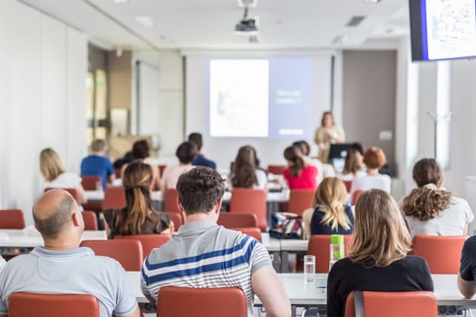 Female speaker giving presentation in lecture hall at university workshop. Audience in conference hall. Rear view of unrecognized participant in audience. Scientific conference event.
