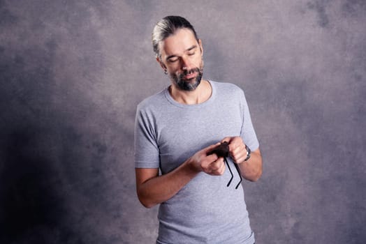 young bearded man ceaning his glasses in front of gray background