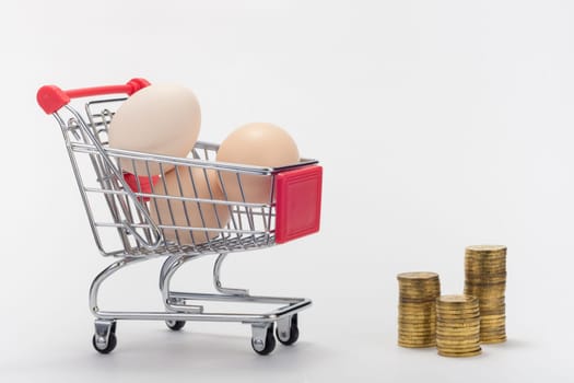 Grocery cart with eggs, next to it are stacks of coins, on a white background