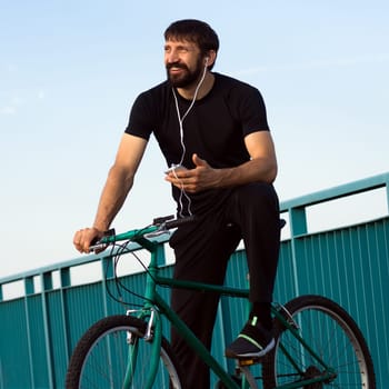 The cheerful smiling cyclist with earphones is using phone outdoors.