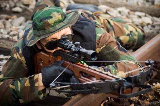 The military man is aiming with crossbow weapon and lying down on railway track.