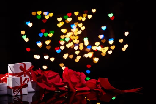 Gift boxes and red roses on a black background with heart-shaped bokeh . Valentine's Day card with copy space. Design element for romantic greeting card, wedding invitation