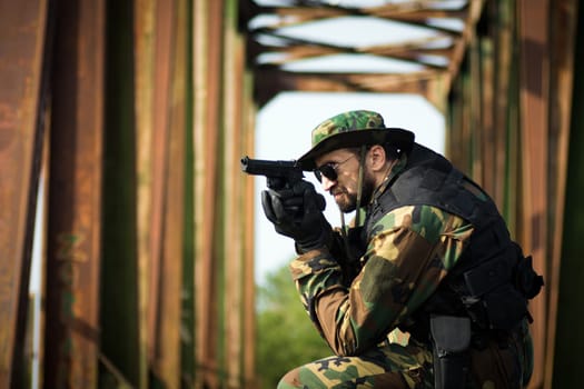 The army soldier in military uniform is aiming with pistol outdoors on old bridge.