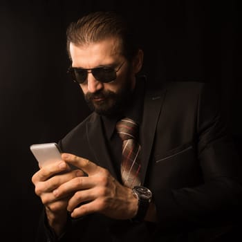 The elegant businessman in black suit is using phone on black background.