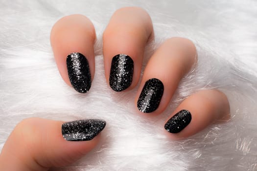 The female hand with black glittered nails is on white furry background.
