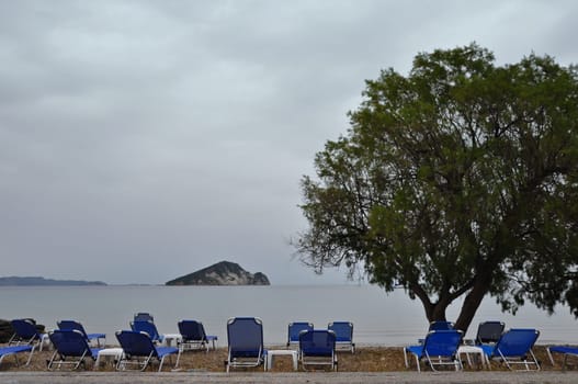 Lounge sunbed chairs with sea view tamarix tree and distant islet, Keri beach, Zakynthos Greece.