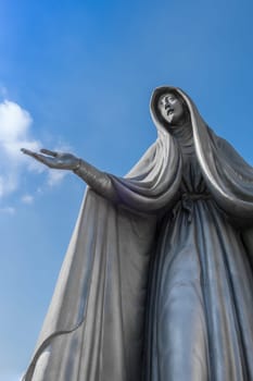 Statue of Virgin Mary in bronze, in a blue sky background with white clouds, with outstretched arms.