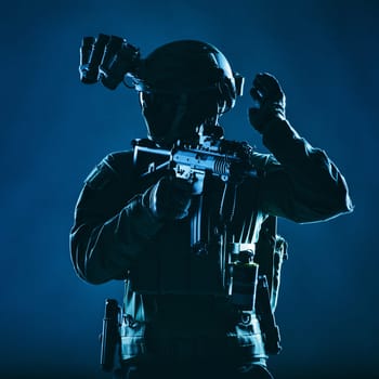 Special operations soldier, SWAT team fighter in mask and glasses, equipped night vision device, armed short barrel service rifle, looking back and showing freeze hand signal, low key studio shoot