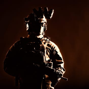Army elite forces member, modern infantryman with hidden face, in tactical ammunition, equipped radio headset, night vision device mounted on helmet, standing with short barrel service rifle in hands