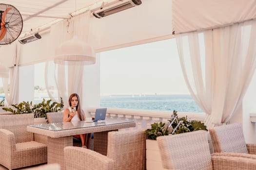 Woman coffee cafe laptop sea. Female freelancer using laptop on coffee break in modern cafe with sea view in the morning.