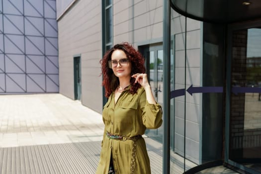 Portrait of a woman standing near a supermarket building. Caucasian model with long brunette hair, wears sunglasses and a khaki dress