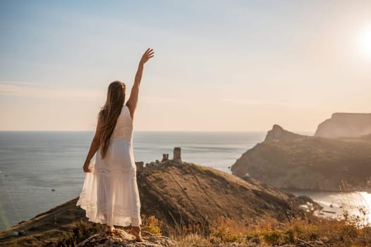 A woman in a white dress stands on a rocky hill overlooking the ocean. She is smiling and she is happy