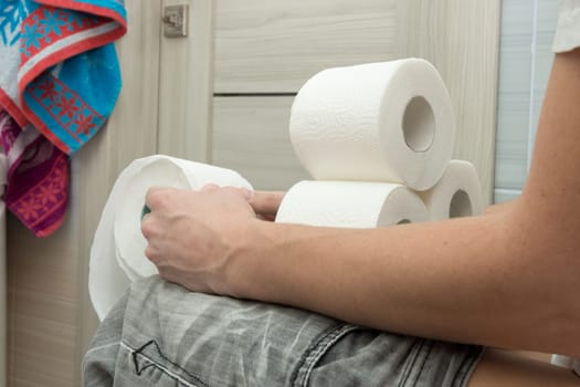 Adult man sitting on the toilet and holding a few rolls of toilet paper