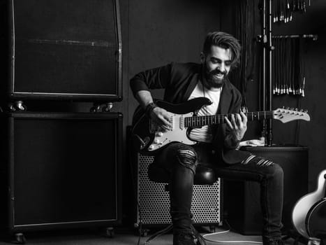 Black and white photo of a man with beard sitting and playing his electric guitar in a recording studio.