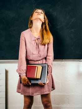 Photo of a frustrated young student holding books in front of a blackboard in class.