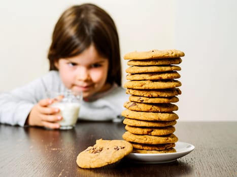 Photo of a young girl staring at a tall pile of chocolate chip cookies while holding a glass of milk. Focus on the cookies.