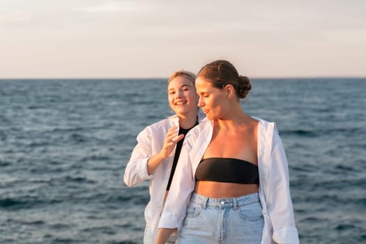 Two women, wearing white shirts, stand on a beach by the ocean. One woman wears a black bikini top and smiles at the camera. The other woman has a hand on her friend's shoulder and looks at her.