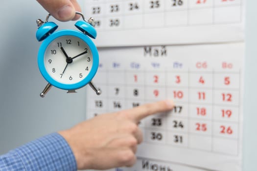 A hand holds a small alarm clock, in the background a hand points to long weekends and holidays on a wall calendar