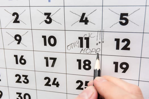 The hand with a pencil writes the words vacation and sea on a wall calendar