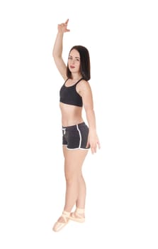 Slim young woman standing in profile in her ballet shoes and one hand.raised, in shorts and black top, isolated for white background.