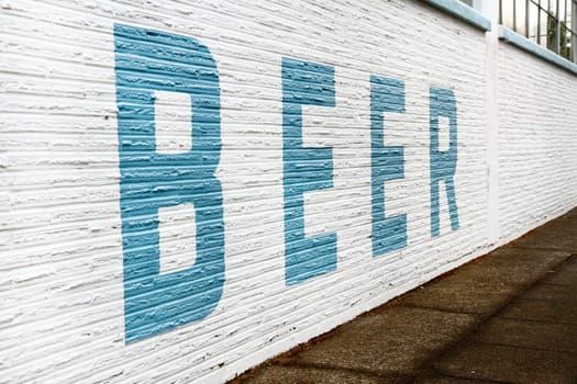 A giant blue letter painted sign spelling BEER on a white brick wall next to a sidewalk.