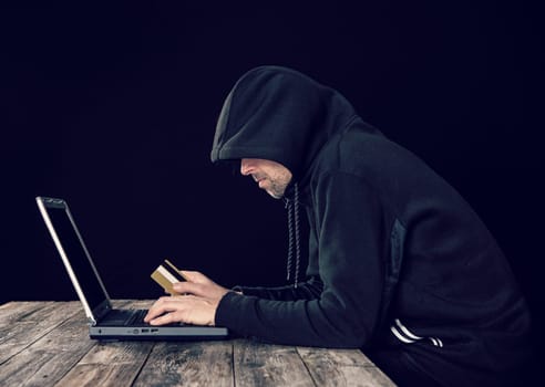 hacker in black hoody with laptop and bank card  in front of black background