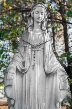 Virgin Mary Statue with open arms. On defocused blurry background a blue sky and green trees. Ideal for concepts and festivity.