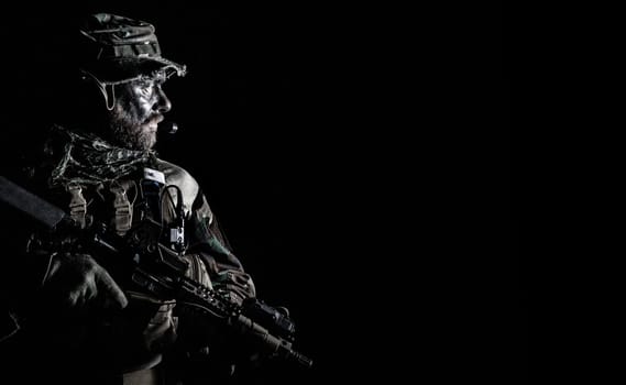 Bearded Special forces United States in Camouflage Uniforms studio shot half length black background. Holding weapons, wearing jungle hat, Shemagh scarf, he is ready to kill. Backlit