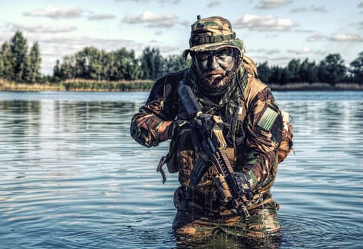 Bearded soldier of special forces in action during river raid in the jungle terrain. He is waist deep in the water and mud and ready to meet enemy, survive and fight in agressive hostile environment