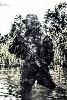 Bearded soldier of special forces in action during river raid in the jungle terrain. He is waist deep in the water and mud and ready to meet enemy, survive and fight in agressive hostile environment