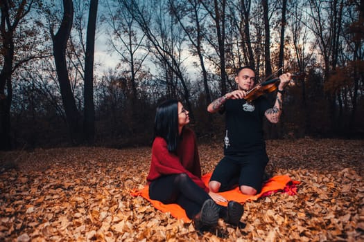 young man and young woman are sitting on a plaid in an autumn forest.