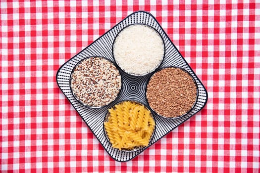 Vegetable food. A decorative plate with four bowls with buckwheat, noodles, rice and quinoa on a red checkered tablecloth. Healthy eating. Top view with space for design.