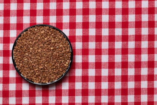 Vegetable food. Macro of a bowl of uncooked raw brown buckwheat grains on a red checkered tablecloth. Healthy eating. Top view with space for product montage.