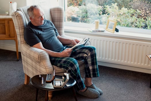 Senior diabetic man is relaxing in the living room of his home with a book. His blood glucose testing kit is on the table next to him.