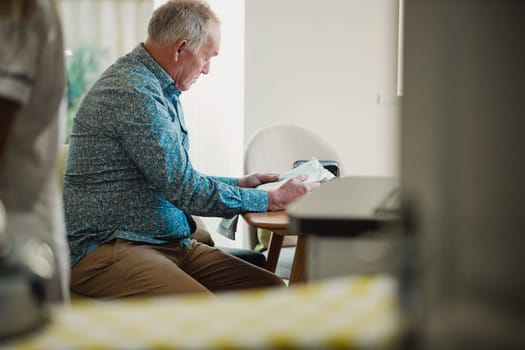 Senior diabetic man is sitting at a dining table reading a newspaper while his carer prepares his medication.