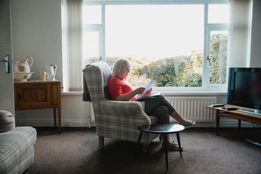 One mature woman is sitting in an armchair in the living room of her home, reading through letters.