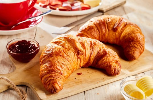 Two fresh baked croissants on wooden cutting board, served with jam and butter in small glass bowls